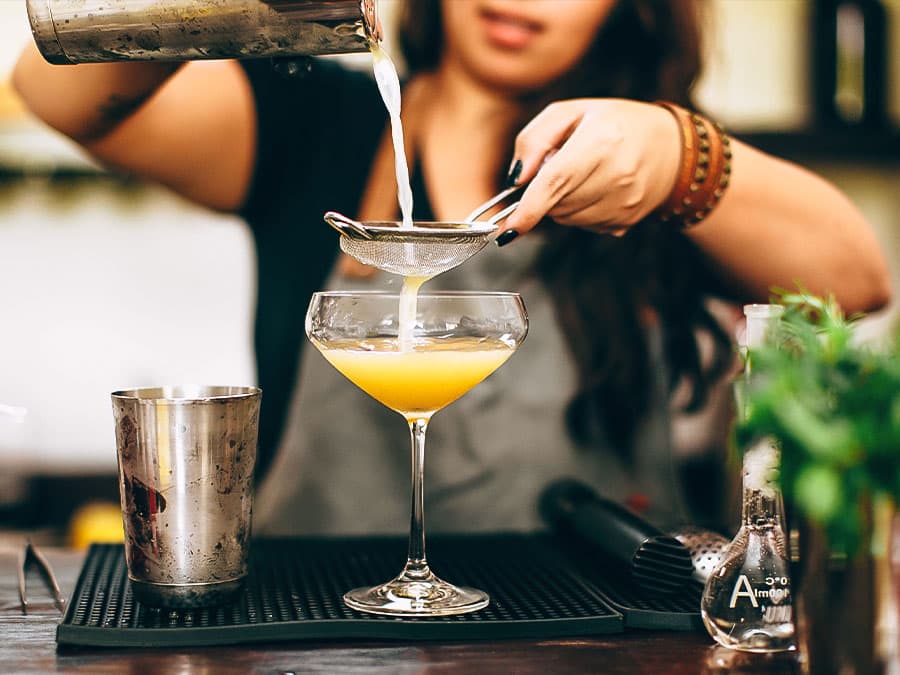 Photo of a bartender pouring juice through a fine-mesh strainer into a coupe glass..